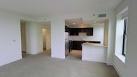 This single-floor space is designed with an open-concept layout that integrates the kitchen and living area into one cohesive, functional environment. The kitchen is appointed with dark espresso cabinetry that contrasts sharply against white walls and a light-toned tile backsplash. Stainless steel appliances, including a refrigerator and range, add a modern touch, while the light granite countertops provide ample prep space. A peninsula breakfast bar extends from the kitchen, offering additional counter space and serving as a natural divider between the kitchen and the open living area. Recessed lighting in the kitchen ensures well-lit task areas. The living area features neutral-toned carpeting and smooth white walls, creating a clean, versatile canvas. A hallway visible from the main living space leads to additional areas of the unit, suggesting a practical and well-organized floor plan. The overall ambiance is contemporary and minimalist, with a neutral palette throughout that allows for flexible interior styling. The space benefits from a simple, uncluttered design that emphasizes functionality and ease of living.