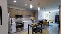 This thoughtfully designed single-floor space showcases a seamless open-concept layout that integrates the kitchen, dining, and living areas into one cohesive and functional environment. The kitchen is appointed with stainless steel appliances, including a range, microwave, and dishwasher, complemented by light wood cabinetry, crisp white lower cabinets, and a stylish mosaic tile backsplash. White quartz-style countertops provide ample prep space, while pendant lighting above adds a warm, modern ambiance. A compact dining area sits adjacent to the kitchen, featuring a dark-toned table with bar-height seating — an efficient and contemporary arrangement ideal for everyday dining. The living area flows naturally from the dining space and is bathed in natural light streaming through large windows, creating a bright and welcoming atmosphere. Neutral tones throughout the space are accented by pops of color in the furnishings, including a mustard yellow accent chair and vibrant wall art. Recessed lighting and a track lighting system ensure well-distributed illumination across the entire open floor plan. The overall aesthetic blends clean lines and modern materials to create a stylish yet highly livable environment.