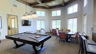 This single-floor amenity space combines comfort and entertainment in a well-appointed, open layout. The room's most striking architectural feature is its coffered ceiling with dark wood beams, lending a refined, traditional character to the space. Tall, multi-pane windows line the walls at two levels, flooding the interior with generous natural light and offering views of the surrounding greenery outside.

At the center of the room stands a full-size pool table, illuminated by a wrought-iron pendant light fixture with classic globe shades, making it the natural focal point for social gatherings. A wall-mounted flat-screen television is positioned nearby, complemented by casual lounge seating upholstered in neutral tones with accent pillows, creating a relaxed viewing area.

For those seeking a more structured setting, the space includes multiple round and rectangular dining tables with cushioned chairs in warm terracotta tones, ideal for casual meals, meetings, or social events. The patterned carpeted flooring ties the various zones together cohesively. Overall, the space strikes a balance between elegance and functionality, offering a versatile environment suited for both active recreation and relaxed socializing.