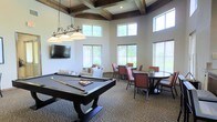 This single-floor amenity space combines comfort and entertainment in a well-appointed, open layout. The room's most striking architectural feature is its coffered ceiling with dark wood beams, lending a refined, traditional character to the space. Tall, multi-pane windows line the walls at two levels, flooding the interior with generous natural light and offering views of the surrounding greenery outside.

At the center of the room stands a full-size pool table, illuminated by a wrought-iron pendant light fixture with classic globe shades, making it the natural focal point for social gatherings. A wall-mounted flat-screen television is positioned nearby, complemented by casual lounge seating upholstered in neutral tones with accent pillows, creating a relaxed viewing area.

For those seeking a more structured setting, the space includes multiple round and rectangular dining tables with cushioned chairs in warm terracotta tones, ideal for casual meals, meetings, or social events. The patterned carpeted flooring ties the various zones together cohesively. Overall, the space strikes a balance between elegance and functionality, offering a versatile environment suited for both active recreation and relaxed socializing.