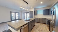 This single-floor residence showcases a contemporary design with a cohesive color palette of gray, white, and warm wood tones. The open-concept kitchen serves as the heart of the space, centered around a generous granite-topped island with an undermount sink, pendant lighting featuring geometric cage-style fixtures, and a stainless steel dishwasher. Gray shaker-style cabinetry lines the perimeter walls, complemented by matching granite countertops, a subway tile backsplash, and a full suite of stainless steel appliances including a refrigerator, range, and microwave. Light wood-look flooring flows throughout the kitchen and living areas, enhancing the bright, airy atmosphere. French doors provide access to an outdoor balcony, allowing natural light to fill the main living space.

The primary bathroom features a dual-sink vanity with granite countertops, gray cabinetry, a large mirror, and a glass-enclosed shower. A secondary bathroom includes a soaking tub, single-sink vanity with matching finishes, and access to a walk-in closet with built-in shelving. Bedrooms are finished with plush carpet and ceiling fans, with windows offering outdoor views. The entry corridor transitions seamlessly into the main living area, completing this well-appointed layout.
