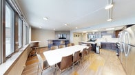 This single-floor communal space is thoughtfully designed to accommodate a variety of social and functional needs. At its center sits a large, elongated white dining table surrounded by modern wooden chairs, providing ample seating for group meals or meetings. The flooring features a mixed-material design combining carpet and hardwood-style tiles, adding visual warmth and texture throughout.

Toward the back of the room, a lounge area includes a raised bar with high stools and a wall-mounted flat-screen television set against a decorative tiled accent wall, creating an inviting space for casual gatherings. Recessed ceiling lighting ensures the area is well-lit throughout.

Adjacent to the lounge, a fully equipped kitchen features dark cabinetry, stainless steel appliances including a range and microwave, a white countertop island with additional bar seating, and a subway tile backsplash. The kitchen is well-appointed for catering or communal cooking. The overall ambiance is clean, modern, and functional, making this space well-suited for both everyday use and hosted events.