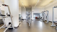 This generously sized fitness facility offers a thoughtfully designed exercise environment spread across a single open floor. The space features a clean, modern aesthetic defined by crisp white walls, durable rubber flooring, and a suspended tile ceiling fitted with linear LED lighting that ensures consistent, bright illumination throughout.

The facility is divided into distinct functional zones. The strength training area is well-appointed with a multi-station cable machine, a Smith machine with an integrated power rack, adjustable benches, and a dedicated free weights section complete with a dumbbell rack and wall-mounted mirrors for form monitoring. The cardio zone houses a row of treadmills and a stationary bike, all positioned along windowed walls that allow ample natural light to flow in and offer views of the surrounding urban skyline.

High clerestory windows in select areas amplify the sense of openness, creating an airy, energizing atmosphere. Full-length mirrors are strategically placed to visually expand the space and support safe training practices. The generous floor area provides comfortable spacing between equipment, allowing multiple users to train simultaneously without crowding. Overall, the facility presents a professional, well-maintained environment suitable for a wide range of fitness activities.