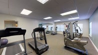 This single-level fitness room offers a comprehensive exercise environment designed to accommodate a variety of workout routines. The space is finished with neutral-toned carpet flooring and features a drop ceiling outfitted with recessed fluorescent panel lighting that ensures consistent, bright illumination throughout. The walls are painted in a combination of neutral beige and sage green tones, contributing to a calm, focused atmosphere.

The room is equipped with an array of cardio machines, including treadmills, elliptical trainers, and a stationary upright bike, all positioned to allow comfortable spacing between users. A multi-station strength training machine provides options for resistance-based workouts, while a medicine ball adds to the variety of available fitness tools. Instructional exercise posters are mounted on the walls for user reference.

Large wall mirrors span one side of the room, visually expanding the space and allowing users to monitor their form during workouts. Windows along the perimeter allow natural light to supplement the overhead lighting. A sanitation dispenser is conveniently mounted near the entrance, promoting hygiene. The room is accessed through a standard door and is well-organized, providing an efficient and functional fitness facility suitable for a range of fitness levels.
