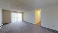 This single-floor unit offers two bedrooms and two bathrooms across a thoughtfully arranged floor plan. The main living area is generously sized, featuring wall-to-wall carpet in a neutral gray tone that extends throughout the space, creating a cohesive and understated aesthetic. A large sliding glass door with vertical blinds dominates one wall of the living area, allowing abundant natural light to fill the room while providing access to an outdoor area. The textured ceiling adds a classic architectural detail consistent throughout the unit. The living space flows naturally into a hallway that connects to the rest of the unit, including the bedrooms and bathrooms. The walls are painted a clean, crisp white, contributing to a bright and open ambiance. The layout is straightforward and functional, offering clear sightlines from the main living area to adjoining spaces. The neutral finishes and open arrangement provide a versatile canvas adaptable to a variety of furnishing styles and personal preferences, making it a practical and comfortable residential space.