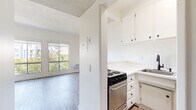 Located in the heart of Westwood, Los Angeles, this 2-bedroom, 1-bathroom unit at 1450 Midvale Ave offers a compact, efficiently designed layout spread across a single floor. The space features light-toned hardwood-style flooring throughout, creating a clean and cohesive aesthetic.

The primary bedroom is the largest room in the unit, offering generous proportions relative to the overall footprint. It is brightened by a pair of large windows that frame treetop and city skyline views, while a wall-unit air conditioner provides climate control. A sliding mirrored closet door adds depth to the room and provides practical storage with built-in shelving and hanging space.

The kitchen is thoughtfully arranged with white cabinetry above and below white countertops, a stainless steel single-basin sink with a matte black faucet, a stainless steel electric range, and a stainless steel refrigerator — maximizing functionality within the compact space.

The bathroom is finished in an all-white palette, featuring a tub/shower combination with tile surround, a single vanity with white countertop, and wood-look tile flooring that ties into the rest of the unit's design language.

A hallway connects the rooms and provides flow throughout the unit. Textured ceilings are present throughout, and the overall light, neutral color scheme keeps the space feeling bright and airy.