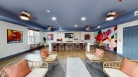 This stylish communal lounge space is thoughtfully designed to encourage gathering and socializing. The interior is anchored by a long, white quartz bar island with modern bar seating, set against a textured accent wall housing a wall-mounted TV, sink, and built-in appliances — ideal for hosting and entertaining. The deep navy-painted ceiling adds a bold design element that contrasts beautifully with the white walls and light wood-look flooring throughout.

The space is enriched by a curated collection of vibrant, large-scale artwork displayed on the walls, including a striking geometric mural in multicolored tones and several vivid canvas paintings that bring energy and personality to the room. Rattan lounge chairs with coral accent pillows, bistro-style tables, and mid-century modern accent chairs are arranged throughout, creating a variety of intimate seating areas suited for both casual conversation and focused work.

Natural light pours in through generous windows and multiple sets of sliding glass doors, which open directly onto an outdoor patio area. Lush tropical potted plants accent the transitions between indoor and outdoor spaces, contributing to the lounge's welcoming, resort-like ambiance. Ceiling fans and recessed lighting ensure comfort throughout the day and evening, making this a versatile and inviting communal retreat.