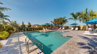 This single-level outdoor amenity space centers around a generously sized swimming pool with clear, blue water, designed for relaxation and recreation. The pool is framed by a well-maintained paver deck that provides ample room for lounging, with numerous chaise lounge chairs arranged around the perimeter for comfort and convenience. Vibrant blue market umbrellas are stationed throughout the deck, offering shaded retreats from the sun. Lush tropical landscaping, including tall palm trees and mature greenery, borders the space, creating a resort-like atmosphere that feels both private and inviting. The surrounding environment hints at a warm, sunny climate — consistent with a Florida setting — with wide-open skies that fill the area with abundant natural light. The overall ambiance is bright, clean, and leisurely, evoking the feel of a premium community pool facility. Additional interior spaces captured in the tour appear to be utility or transitional areas. The outdoor pool deck serves as the standout feature of this space, offering a well-appointed environment ideal for outdoor recreation and relaxation.
