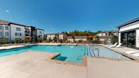 This outdoor amenity area is centered around a generously sized swimming pool with crystal-clear blue water, complemented by a separate spa. The pool deck is finished in smooth concrete and furnished with multiple cushioned lounge chairs and tables, offering ample space for sunbathing and relaxation. Vibrant orange market umbrellas are positioned throughout the deck, providing shaded spots for residents. A wrought iron perimeter fence encloses the pool area, ensuring privacy and security.

Adjacent to the pool, a covered outdoor corridor connects key amenity spaces, including a 24-hour fitness center with glass-panel entry doors. An open-air cabana lounge area features ceiling fans, sheer curtains, and comfortable seating, creating a semi-private retreat. The surrounding landscape includes mature pine trees and manicured plantings, adding a natural backdrop to the modern architectural aesthetic of the multi-story residential buildings visible in the background. The overall ambiance is bright and resort-like, designed to maximize outdoor living under sunny skies.