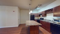 This single-floor residential unit offers a thoughtfully designed open-concept layout that seamlessly connects the living area and kitchen. The kitchen is a standout feature, appointed with rich dark wood cabinetry, granite-style countertops, a central island with pendant lighting, track lighting, black appliances including a refrigerator and gas range, a built-in dishwasher, microwave, and an upper wine rack, adding a refined touch to the space. Hardwood-style flooring runs throughout the main living areas, complementing the neutral, warm-toned walls. The living area is generously sized with a ceiling fan and receives natural light through a transom window near the entryway, creating a bright and welcoming atmosphere. Two bedrooms are carpeted for comfort, each equipped with ceiling fans and windows fitted with blinds that allow for natural light and privacy. The two bathrooms mirror each other in design, each featuring dark wood vanities with granite-style countertops, tub-shower combinations with white tile surrounds, and hardwood-style flooring. Clean lines, consistent finishes, and a cohesive color palette give the entire unit a polished and functional character.