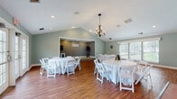 Located at 1331 Trinity Pl. in Middletown, Ohio, this single-floor event venue offers a well-appointed space designed to host a variety of gatherings. The main hall is the centerpiece of the venue, featuring soaring vaulted ceilings that lend an open, airy feel to the room. Warm hardwood flooring runs throughout, complementing the soft sage-green walls and recessed lighting. An elegant chandelier adds a touch of sophistication overhead. The hall is configured with round banquet tables and white folding chairs, offering comfortable seating for guests. A raised stage at one end of the room is flanked by speaker stands, making it well-suited for live performances, presentations, or ceremonies. Large French doors along one wall invite abundant natural light and provide access to the exterior. A dedicated bar and kitchen area is conveniently positioned adjacent to the main hall. The kitchen features dark cabinetry, black appliances including a refrigerator, range, and microwave, and a generous light-colored countertop. Terra cotta tile flooring defines this service area. A wide pass-through counter connects the kitchen to the main hall, facilitating efficient food and beverage service. The overall ambiance is clean, bright, and versatile, making this venue a practical and attractive choice for events of all kinds.