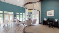 This leasing office makes a striking first impression with its dramatic vaulted ceilings and bold teal accent walls that contrast crisply against bright white surfaces. A standout architectural detail is the sweeping curved wooden archway that elegantly frames the transition between the welcoming lounge area and the dedicated workstation zone, adding warmth and visual interest throughout the space.

The main lounge area is thoughtfully appointed with modern mid-century style seating, including plush tufted chairs and a tufted sofa arranged around a glass-top coffee table atop a patterned area rug. A decorative fireplace serves as a focal point, flanked by large windows and French doors that invite an abundance of natural light and offer views of the surrounding outdoor deck and greenery.

Adjacent to the lounge, a casual dining and meeting area provides bistro-style tables and chairs, bathed in light streaming through a full wall of windows and French doors leading to the exterior. The workstation area, subtly set apart by the curved archway and a raised carpeted platform, houses multiple staff desks equipped for day-to-day leasing operations, accented with lush indoor plants.

Throughout, wood-look flooring anchors the space with a contemporary yet inviting feel, while curated artwork and coordinated teal and yellow accent pieces unify the design, creating a professional yet comfortable environment for prospective residents.