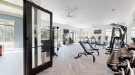 Located at 350 Bay Laurel Dr in Saint Augustine, FL, this single-floor fitness center offers a comprehensive workout facility across two distinct exercise rooms. The main exercise area is expansive and bright, featuring soaring ceilings, large black-framed floor-to-ceiling windows, and herringbone-patterned carpet tile flooring. This primary space is outfitted with a full array of strength machines, a dumbbell rack, cable systems, treadmills, stair climbers, stationary bikes, and a rowing machine. Oversized ceiling fans and wall-mounted TVs are distributed throughout, while a motivational typography mural serves as a striking accent wall. A full-length mirror along one wall enhances the sense of space. Natural light pours in from multiple directions, with views of lush Florida greenery providing an energizing backdrop. The second exercise room offers a distinct functional training environment, featuring light hardwood flooring, soft teal-blue walls, and coffered ceiling details. This room is equipped with a TAC wall-mounted rig holding kettlebells, medicine balls, resistance bands, and weight plates, alongside a heavy punching bag and plyo boxes. A large mirror wall and wall-mounted TV complete the space, making it ideal for group classes or individual functional training sessions.