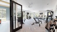 Located at 350 Bay Laurel Dr in Saint Augustine, FL, this single-floor fitness center offers a comprehensive workout facility across two distinct exercise rooms. The main exercise area is expansive and bright, featuring soaring ceilings, large black-framed floor-to-ceiling windows, and herringbone-patterned carpet tile flooring. This primary space is outfitted with a full array of strength machines, a dumbbell rack, cable systems, treadmills, stair climbers, stationary bikes, and a rowing machine. Oversized ceiling fans and wall-mounted TVs are distributed throughout, while a motivational typography mural serves as a striking accent wall. A full-length mirror along one wall enhances the sense of space. Natural light pours in from multiple directions, with views of lush Florida greenery providing an energizing backdrop. The second exercise room offers a distinct functional training environment, featuring light hardwood flooring, soft teal-blue walls, and coffered ceiling details. This room is equipped with a TAC wall-mounted rig holding kettlebells, medicine balls, resistance bands, and weight plates, alongside a heavy punching bag and plyo boxes. A large mirror wall and wall-mounted TV complete the space, making it ideal for group classes or individual functional training sessions.