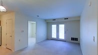 This single-floor space presents a clean, understated interior designed for comfortable everyday living. The main living area is carpeted in a neutral tone, creating a soft and cohesive foundation throughout the room. A prominent pair of French doors dominates one wall, allowing natural light to filter in and providing a potential connection to an outdoor area. The doors are framed in white, complementing the light-colored walls and trim that give the space a bright, airy feel. Ceiling-mounted HVAC vents are evenly distributed, ensuring consistent climate control throughout. A thermostat panel is visible on one wall, adding a practical touch to the otherwise minimal decor. An adjacent room is accessible through a wide interior doorway, also carpeted and finished with matching white walls, suggesting a similarly open and versatile space. The overall layout feels open and well-proportioned, with smooth ceilings and simple architectural lines that keep the interior feeling uncluttered. The neutral palette and consistent finishes throughout give the space a functional yet inviting character suitable for a variety of uses.