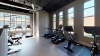 Located at 2300 N Davidson St in Charlotte's NoDa neighborhood, this single-floor fitness center offers a well-equipped and thoughtfully designed workout environment. The space features an open layout with an exposed, dark-painted ceiling that lends an industrial character, while large windows along multiple walls flood the interior with natural light and provide views of the surrounding outdoor area, including a pool and courtyard.

The cardio zone is stocked with TRUE brand treadmalls, ellipticals, stair climbers, and stationary spin bikes, each positioned to take advantage of the natural light streaming through the generous windows. A ceiling fan adds comfort to this section of the space.

The strength training area is equally comprehensive, housing a multi-functional cable and power rack system, adjustable benches, a full dumbbell rack, medicine balls, stability balls, and additional cable machines. The durable rubber and tile flooring throughout supports a range of workout activities safely.

Built-in shelving and cabinetry near the entrance provide storage for accessories and personal items, and glass double doors connect the space to adjacent amenities. The overall ambiance is clean, modern, and motivating, with neutral gray walls and ample equipment density that supports a full range of fitness pursuits.