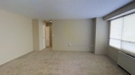 This single-floor space offers a clean and functional layout designed for comfortable everyday living. The main living area is spacious and open, featuring wall-to-wall carpeting in a warm beige tone that adds softness underfoot and contributes to a quiet, understated ambiance throughout. The walls are finished in a neutral tan hue, creating a versatile backdrop that adapts well to a variety of furnishing styles and décor choices. Recessed lighting in the ceiling provides focused illumination, keeping the space feeling bright and well-lit. A hallway extends from the main area, visible in the background, leading to additional rooms and suggesting a practical, connected floor plan. The hallway is finished consistently with the rest of the space, maintaining the overall cohesive aesthetic. Utility provisions, including an electrical outlet and a wall-mounted heating or ventilation panel, are thoughtfully integrated into the walls, ensuring the space is well-equipped for daily use. The overall design is straightforward and low-maintenance, making it a practical and adaptable space suited to a range of occupants and uses.