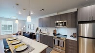 This single-floor residence offers a thoughtfully designed open-concept layout that seamlessly connects the kitchen, dining, and living areas. The kitchen is a standout feature, outfitted with dark wood-grain flat-front cabinetry extending to the ceiling, a crisp white subway tile backsplash, and a full suite of stainless steel appliances including a refrigerator, electric range, built-in microwave, and dishwasher. A white quartz peninsula with bar seating and a pendant light overhead defines the dining space and enhances the kitchen's functionality. Light hardwood-style flooring runs throughout the unit, contributing to a warm, cohesive aesthetic.

The living area is bright and welcoming, benefiting from large windows that invite abundant natural light and views of the surrounding exterior. A private door provides direct outdoor access from this space. The bathroom is well-appointed with dark wood-grain cabinetry, an above-counter sink with a white vanity top, an overhead medicine cabinet, and a tiled tub-shower combination. Recessed and flush-mount lighting throughout the unit maintains a clean, modern ambiance. Overall, the space presents a contemporary design with a neutral palette and quality finishes.