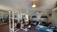 This fitness center is a single-floor facility designed to support a comprehensive workout experience. The main exercise area is finished with durable, dark carpeting suited for gym use and is illuminated by flush-mount ceiling fixtures that provide consistent overhead lighting throughout the space. A large wall of mirrors along one side of the room visually expands the area and allows users to monitor their form during exercise.

The cardio section is well-appointed, featuring multiple Precor treadmills, an elliptical trainer, and a recumbent stationary bike, offering a variety of low-impact and high-intensity aerobic options. The strength training area includes a multi-function cable and weight machine paired with adjustable benches, accommodating a wide range of resistance exercises.

The facility benefits from natural light streaming through a window fitted with horizontal blinds and a set of glass-paned French doors, which also provide direct access to the exterior. Climate comfort is maintained by a wall-mounted mini-split HVAC unit. The textured ceiling and neutral gray walls give the space a clean, no-frills aesthetic. A tiled entryway near the French doors creates a practical transition zone, and a restroom is accessible on-site, adding convenience for users during their workouts.
