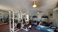 This fitness center is a single-floor facility designed to support a comprehensive workout experience. The main exercise area is finished with durable, dark carpeting suited for gym use and is illuminated by flush-mount ceiling fixtures that provide consistent overhead lighting throughout the space. A large wall of mirrors along one side of the room visually expands the area and allows users to monitor their form during exercise.

The cardio section is well-appointed, featuring multiple Precor treadmills, an elliptical trainer, and a recumbent stationary bike, offering a variety of low-impact and high-intensity aerobic options. The strength training area includes a multi-function cable and weight machine paired with adjustable benches, accommodating a wide range of resistance exercises.

The facility benefits from natural light streaming through a window fitted with horizontal blinds and a set of glass-paned French doors, which also provide direct access to the exterior. Climate comfort is maintained by a wall-mounted mini-split HVAC unit. The textured ceiling and neutral gray walls give the space a clean, no-frills aesthetic. A tiled entryway near the French doors creates a practical transition zone, and a restroom is accessible on-site, adding convenience for users during their workouts.