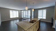 This single-floor space showcases a thoughtfully designed open-concept layout that seamlessly connects the kitchen and living areas. The kitchen serves as the centerpiece, anchored by a generously sized island topped with elegant speckled granite countertops. The island features a double basin stainless steel sink and ample cabinetry finished in crisp white with brushed nickel hardware, providing both style and functionality. Two cylindrical pendant lights hang above the island, casting warm, focused illumination over the workspace.

The open floor plan flows naturally into the adjoining living area, all unified by rich, dark hardwood flooring that adds warmth and sophistication throughout. The walls are finished in a soft, neutral gray tone that complements the overall palette and creates a calm, modern ambiance.

Natural light is a defining feature of this space, with multiple large windows fitted with white horizontal blinds lining the walls of both the kitchen and living areas. These windows invite abundant daylight, enhancing the sense of openness and airiness. Recessed ceiling lighting supplements the natural light, ensuring the space remains bright and well-lit at all times. The overall design is clean, contemporary, and highly functional, making it an inviting environment for both everyday living and entertaining.