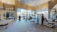 This single-floor fitness center is thoughtfully designed to deliver a premium workout experience in a spacious, inviting environment. The facility boasts dramatically high ceilings adorned with striking decorative wood slat accents and contemporary circular pendant lighting, creating an energetic yet refined atmosphere. Large floor-to-ceiling windows line one wall, flooding the space with abundant natural light and offering open views of the surrounding exterior. The flooring throughout features a durable, patterned commercial carpet tile in neutral tones that adds visual texture while supporting heavy equipment use. The strength training area is comprehensively appointed with a full rack of dumbbells, a power rack with barbell, functional cable machines, and a variety of resistance and weight stations suitable for all fitness levels. Floor-to-ceiling mirrors with an elegant arched detail visually expand the space and allow users to monitor their form during workouts. A large wall-mounted flat-screen television keeps members entertained and informed. The cardio section is lined with multiple treadmills, rowing machines, and additional aerobic equipment positioned to take advantage of the natural light from the expansive windows. Recessed lighting complements the pendant fixtures overhead, ensuring consistent illumination throughout. A water station and additional amenities are conveniently integrated into the layout, rounding out this polished, fully equipped fitness facility.