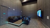 Located in Bradenton, FL, this single-floor amenity space is designed as a premium media and entertainment lounge. The room makes an immediate impression with its soaring ceilings and richly textured, dark charcoal walls that lend the space a dramatic, immersive atmosphere reminiscent of a private cinema. The flooring is covered in a neutral, patterned carpet that complements the dark tonal palette throughout.

The seating area is thoughtfully arranged with multiple deep, dark leather sofas and individual club chairs, all accented with teal and blue throw pillows that add a refined pop of color. A raised platform section with subtle LED accent lighting beneath creates a tiered viewing arrangement, directing attention toward the large wall-mounted flat-screen television positioned at the front of the room. In-wall speakers are integrated seamlessly into the design, reinforcing the dedicated media experience.

Toward the rear of the space, a separate dining or meeting area features a dark wood table surrounded by upholstered chairs, accompanied by a pair of abstract teal-accented wall art pieces that brighten the corner. A small illuminated accent piece on the table adds a warm, ambient glow. The overall design is cohesive, moody, and modern, making this space well-suited for private screenings, group gatherings, or relaxed entertainment in a polished, upscale setting.