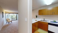 This single-floor residence offers a thoughtfully organized layout that balances open living with functional, well-defined spaces. The living area is generously proportioned, finished with plush wall-to-wall carpeting and neutral warm tones that create a welcoming ambiance. A ceiling fan adds comfort overhead, while large sliding glass doors open to a private balcony overlooking mature trees, flooding the room with natural light and greenery views.

The kitchen is efficiently designed in an L-shaped configuration, featuring warm wood-grain cabinetry extending to the upper walls, white countertops, a stainless steel sink, a dishwasher, and an electric range. Durable hardwood-style flooring grounds the kitchen and dining area, which transitions smoothly into the carpeted living space.

A dedicated vanity area provides a practical and elegant grooming station, complete with a floating white shelf, a single drawer, and a large wall-mounted mirror — positioned conveniently near a closet.

The bathroom features a wide white vanity with generous counter space, ample under-sink storage, a wall mirror, and warm tile flooring, offering both functionality and a clean, classic aesthetic.

Throughout the residence, soft neutral wall colors, consistent flooring choices, and abundant natural light from multiple sliding glass door access points contribute to a cohesive and comfortable atmosphere.