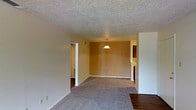 This single-floor space offers a straightforward and functional layout designed for comfortable everyday living. The main living and dining area is an open, connected space highlighted by warm golden-tan walls that create an inviting ambiance. Plush wall-to-wall carpeting covers much of the floor, offering comfort underfoot, while hardwood-style flooring is visible near the entryway, adding a contrasting design touch. A classic textured popcorn ceiling runs throughout, and a simple pendant light fixture hangs centrally over the dining area, providing focused illumination. The open layout allows for flexible furniture arrangement, making it easy to define distinct living and dining zones within the shared space. Doorways lead to additional areas of the home, including what appears to be a hallway providing access to other rooms. The clean white trim and baseboards offer a crisp contrast against the warm wall tones, while the neutral color palette throughout keeps the space feeling cohesive and versatile. Overall, the space presents a practical and welcoming environment with a no-frills design that emphasizes functionality and ease of living.