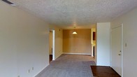 This single-floor space offers a straightforward and functional layout designed for comfortable everyday living. The main living and dining area is an open, connected space highlighted by warm golden-tan walls that create an inviting ambiance. Plush wall-to-wall carpeting covers much of the floor, offering comfort underfoot, while hardwood-style flooring is visible near the entryway, adding a contrasting design touch. A classic textured popcorn ceiling runs throughout, and a simple pendant light fixture hangs centrally over the dining area, providing focused illumination. The open layout allows for flexible furniture arrangement, making it easy to define distinct living and dining zones within the shared space. Doorways lead to additional areas of the home, including what appears to be a hallway providing access to other rooms. The clean white trim and baseboards offer a crisp contrast against the warm wall tones, while the neutral color palette throughout keeps the space feeling cohesive and versatile. Overall, the space presents a practical and welcoming environment with a no-frills design that emphasizes functionality and ease of living.