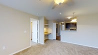 This single-floor residence showcases an open-concept layout that connects the living area and kitchen, creating a spacious and functional flow throughout. The living area is finished with plush wall-to-wall carpet in a neutral tone, complemented by light beige walls and white baseboards that contribute to a clean, cohesive aesthetic. A ceiling fan overhead provides both lighting and air circulation to the main living space.

A built-in desk nook with open shelving and a granite-topped work surface is integrated into the wall, offering a practical workspace without occupying additional floor space. Adjacent to this nook, a white paneled door leads to what appears to be a bathroom or utility area.

The kitchen is partially visible from the living area, separated by a raised breakfast bar counter. Dark espresso cabinetry contrasts with lighter countertops, and a pendant light fixture hangs above the bar, adding a touch of style. Hardwood-style flooring transitions from carpet in the kitchen zone, further defining the two areas. A hallway visible in the background suggests additional rooms beyond the main living space.