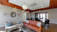 This single-floor space offers a thoughtfully designed interior that blends comfort and functionality in a professional setting. The main lounge area serves as a central gathering space, furnished with a warm terracotta sofa and complementary patterned seating arranged around a glass coffee table. A decorative drum chandelier anchors the room from above, while recessed lighting throughout ensures consistent, well-distributed illumination. The ceiling features a tray detail finished in a rich burnt orange accent, adding architectural depth and a distinctive visual focal point that ties together the warm color palette of the space. Neutral gray walls provide a clean backdrop that balances the bold accents throughout. An open passage leads into a dedicated dining area furnished with dark glass-top tables and black chairs, suitable for meetings or group gatherings. French doors at the rear of the dining area allow natural light to filter in and offer a view to the exterior. A decorative sunburst mirror and framed wall art contribute to the polished, welcoming ambiance. The space also includes a private room accessible through a doorway off the main lounge, providing additional flexibility for various uses. Overall, the layout is open, functional, and professionally appointed.