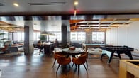This thoughtfully designed single-floor amenity space blends work and leisure seamlessly through a cohesive mid-century modern aesthetic. The open-plan layout is anchored by rich hardwood flooring, exposed concrete columns, and a striking coffered ceiling detailed with warm wood accents and integrated lighting that adds depth and visual interest throughout.

The game and recreation area is a standout feature, equipped with a full-size billiards table and foosball table, set atop a vibrant geometric-patterned carpet. Adjacent to this is a comfortable media lounge with a large wall-mounted flat-screen television, leather seating, and accent chairs arranged around a sculptural coffee table, all bathed in natural light from expansive windows overlooking urban views.

A co-working area provides long communal tables with brass-accented seating beneath dome pendant lights, ideal for productivity. Separate glass-walled conference rooms with folding glass doors offer private meeting spaces while maintaining visual connectivity to the broader floor.

The compact kitchenette features slate-blue cabinetry, clean white countertops, light subway tile walls, and open brass shelving, providing a functional refreshment area. Together, these zones create a dynamic, design-forward environment suited for both collaboration and relaxation.