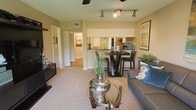 This single-floor residence features an open-concept layout that connects the living, dining, and kitchen areas into one cohesive, flowing space. The living area is finished with plush wall-to-wall carpeting in a warm neutral tone, providing a comfortable foundation for the room. A ceiling fan overhead and a track lighting system with adjustable spotlights ensure flexible, well-distributed illumination throughout the space.

The dining area is anchored by a dark, high-top table paired with matching bar-height chairs, creating a casual yet stylish setting. This transitions naturally into the kitchen, which features white cabinetry, mirrored backsplash accents, and dark countertops, adding a touch of contemporary elegance. The kitchen is equipped with modern appliances and benefits from the reflective surfaces that enhance the sense of brightness.

Neutral beige walls run throughout, providing a warm and cohesive backdrop. A hallway visible in the background suggests additional private spaces beyond the main living area. Overall, the space presents a practical and modern open-plan design with clean lines and a welcoming ambiance.