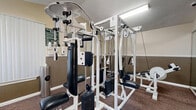 Located in Bradenton, Florida, this dedicated exercise room offers a well-equipped fitness space designed to support a complete workout routine. The room features a two-tone wall treatment, with a warm beige upper half and a darker wainscoting-style lower panel, complemented by plush carpeted flooring in a deep brown tone that adds comfort underfoot.

The space is outfitted with a notable range of fitness equipment, including a treadmill positioned near a window with vertical blinds that allow natural light to filter in. A large multi-station cable machine serves as the centerpiece of the room, offering a variety of strength-training options. Multiple weight benches and leg extension stations round out the strength equipment selection, providing versatility for full-body workouts.

Large wall mirrors line one side of the room, visually expanding the space and allowing users to monitor their form during exercise. Overhead fluorescent lighting ensures the room is well-lit throughout. A door provides direct access to an outdoor area, offering convenient ventilation. The textured ceiling and vaulted angles in portions of the room add subtle architectural character. Overall, this exercise room delivers a practical, functional fitness environment well-suited for daily use.