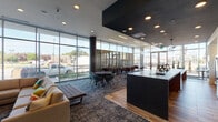 This contemporary single-floor amenity lounge is defined by its expansive floor-to-ceiling curtain-wall glazing, flooding the interior with abundant natural light and offering sweeping views of the surrounding streetscape. The open-plan layout is thoughtfully divided into distinct functional zones that flow seamlessly into one another.

The social and dining area features a generous dark-topped communal bar with leather-upholstered counter seating, accompanied by high-top bistro tables and mixed seating arrangements set atop patterned carpet tiles. A full kitchenette wall anchors this zone, complete with wall-mounted ovens, a beverage station, under-counter refrigeration, and a subway tile backsplash framing a large flat-screen television above. Industrial-style pendant lighting adds character overhead.

The lounge area transitions into a more relaxed setting, furnished with plush sofas, a tufted bench, and mid-century modern accent chairs arranged around a stacked natural stone fireplace with a linear hearth. A teal accent wall showcases bold abstract artwork, lending the space a curated, gallery-like quality. Soft carpet underfoot and warm recessed lighting create an inviting atmosphere throughout.

The interplay of hardwood-look flooring, textured carpeting, dark ceiling planes, and natural stone creates a rich, layered aesthetic that balances contemporary design with comfortable livability.