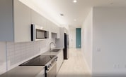 This 600 square foot apartment in Durham, NC, offers a thoughtful layout maximizing space and functionality. The single-floor plan includes one bedroom, measuring 100 square feet, one bathroom, and a hallway connecting the living spaces. The apartment showcases a contemporary design with clean lines and a neutral color palette. Light wood-toned flooring runs throughout, complementing the crisp white walls and creating an open, airy ambiance. Large windows in the main living area provide ample natural light and offer impressive city views, enhancing the sense of space. The bathroom features a modern aesthetic with a sleek glass shower enclosure and white fixtures. The bedroom, while compact, is well-proportioned and includes built-in lighting. Overall, this apartment combines efficient use of space with modern amenities, making it an ideal urban dwelling for those seeking a streamlined lifestyle in the heart of Durham.