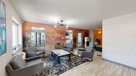 Located at 645 Wolff Street in Denver's vibrant 80204 corridor, this CoWorking Lounge at Alpine Place offers a well-appointed, single-floor shared workspace designed to support both productivity and relaxation. The main lounge area serves as the social hub of the space, anchored by a bold, floor-to-ceiling mountain landscape mural in warm amber tones that adds dramatic visual interest. Modern mid-century-style seating — consisting of grey upholstered chairs arranged around a rectangular coffee table — rests atop a graphic patterned area rug over light wood-look tile flooring. A striking sputnik chandelier provides ambient overhead lighting throughout the lounge. A dedicated workstation nook, finished with a vibrant orange accent wall and dual sconce lighting, accommodates two desktop computer setups with ergonomic seating. The kitchenette features clean white cabinetry, a stainless steel dishwasher and refrigerator, a countertop microwave, coffee station, and a three-dimensional textured tile backsplash, with a window providing natural light. The entry area includes a reception-style desk, whiteboard, and lush green carpet detailing. Glimpses of an on-site fitness area with a treadmill and wall-mounted TV round out this comprehensive community lounge.
