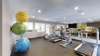 This fitness center offers a dedicated space for residents to pursue their health and wellness goals. The single-floor layout is thoughtfully designed to maximize functionality, featuring durable rubber flooring that provides a safe, supportive surface for a variety of exercises. The room is illuminated by recessed lighting and benefits from natural light streaming in through multiple windows, creating a bright and energizing atmosphere.

The cardio section is well-appointed with a treadmill, elliptical trainer, and a stationary bike, catering to a range of aerobic workout preferences. A large wall-mounted mirror spans one side of the room, enhancing the sense of space and allowing users to monitor their form during workouts. A flat-screen television is mounted on the wall, providing entertainment during exercise sessions.

Complementing the cardio equipment, an assortment of colorful stability and exercise balls adds versatility for core training, stretching, and balance exercises. Natural light from the windows and the open, uncluttered layout contribute to a welcoming and motivating environment. French doors provide easy access to the adjacent area, further integrating the fitness center into the broader community facility. Overall, this space delivers a practical and inviting setting for daily fitness routines.