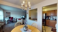 Located at 835 Blooming Grove Turnpike in New Windsor, New York, this single-floor home offers a comfortable and functional layout with a warm, inviting ambiance throughout. The dining area serves as a central hub, featuring a classic brass candelabra chandelier that adds a traditional elegance to the space. A round wooden dining table with matching Windsor-style chairs provides a welcoming setting for everyday meals and gatherings. A partial wall with a pass-through opening connects the dining area to an adjacent space, enhancing the sense of openness and flow between rooms. The living room is visible from the dining area and features neutral-toned furnishings, carpeted flooring, and a television setup, creating a relaxed and cozy retreat. Through the pass-through opening, a secondary room is visible, hinting at additional flexible living space. The kitchen is accessible from the dining area and showcases tile flooring, warm wood cabinetry, and a full-size refrigerator, offering practical functionality. The layout of this home encourages easy movement between living, dining, and kitchen spaces, making it well-suited for comfortable everyday living. Natural and ambient lighting throughout contribute to a bright and welcoming atmosphere across all areas of the home.