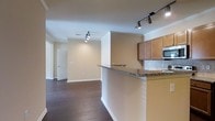 This single-floor residence offers a thoughtfully designed open-concept layout that seamlessly connects the kitchen and living areas, creating a spacious and inviting atmosphere. The kitchen is equipped with warm honey-toned cabinetry, granite countertops, a built-in microwave, and a decorative tile backsplash. A peninsula counter extends from the kitchen, providing both additional prep space and a natural divider between the cooking and living zones. Rich, dark hardwood flooring runs throughout the main living area, adding warmth and sophistication to the space. Crown molding lines the ceilings, lending a polished, refined character to the interior. Track lighting installed on the ceiling offers adjustable, directional illumination, ensuring the space is both functional and well-lit. The neutral warm-beige walls create a versatile backdrop suited to a variety of design styles. A doorway visible at the far end of the living area leads to additional rooms, hinting at a practical and well-organized floor plan. The overall ambiance is clean, modern, and move-in ready, with architectural details that elevate the everyday living experience.