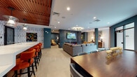 This single-floor communal lounge is thoughtfully designed with distinct zones that cater to a variety of social and relaxation needs. The space is anchored by a large sectional sofa in the central lounge area, accented with bold orange throw pillows that contrast beautifully against deep teal walls. A wall-mounted flat-screen TV sits above a sleek linear fireplace clad in dark metal paneling, creating a dramatic focal point.

Adjacent to the main lounge is a well-appointed bar area featuring a broad white quartz countertop, warm wood slatted ceiling panels, and a row of plush orange velvet bar stools with brass-tipped legs. Open shelving and a herringbone tile backsplash complete the bar's polished aesthetic. Pendant lights in glass shades hang above, adding refined illumination.

A more intimate lounge alcove features tufted velvet seating, a vibrant blue area rug, curated artwork, a decorative sunburst mirror, and a contemporary multi-bulb chandelier overhead. Built-in bookshelves line one wall, adding a library-like warmth. Large glazed doors in the seating area invite natural light and outdoor views. Throughout, light tile flooring, recessed lighting, and eclectic art installations contribute to a cohesive, upscale ambiance.