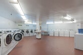 Located at 1850 Rathburn Rd E in Mississauga, Ontario, this well-equipped shared laundry facility spans a generously sized single floor, offering ample room for residents to carry out their laundry needs comfortably. The space is organized across two interconnected rooms, both finished with durable terracotta-toned ceramic tile flooring and clean white walls accented with white tile wainscoting along the lower sections.

The main laundry area houses a large number of front-loading and top-loading washing machines alongside matching dryers, all in white and arranged along the perimeter walls to maximize open floor space. A central folding island and wall-mounted folding surfaces provide convenient areas for sorting and folding laundry. A utility sink with exposed plumbing offers additional functionality for pre-treatment or hand-washing tasks.

The secondary area serves as a waiting and relaxation space, furnished with a black leather sofa and a small round table with seating, making it comfortable for residents who choose to wait during their laundry cycles. A cork bulletin board and a wall clock are mounted near the entrance, adding a practical, community-oriented touch. Bright fluorescent overhead lighting illuminates both rooms evenly, and a fire extinguisher is visibly mounted for safety. The overall layout is clean, functional, and efficiently designed to serve a multi-unit residential building.