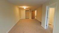 This single-floor residential space offers a clean, functional layout designed for practical living. The main room is generously proportioned, featuring neutral-toned wall-to-wall carpeting that adds warmth and comfort underfoot. The room is illuminated by a ceiling-mounted light fixture with a fan, providing both ambient lighting and air circulation. White painted walls and white baseboards contribute to a bright, clean aesthetic throughout the space.

A notable feature of the room is a large built-in closet with a classic paneled white door, offering substantial storage space complete with a hanging rod. An additional interior door, finished in a warm wood tone, leads to an adjoining area, suggesting a connected layout that may include a bathroom or hallway. The doorway reveals a tiled floor beyond, indicating a transition to a hard-surface area.

The overall design is straightforward and unfussy, with smooth, even walls and consistent flooring throughout the carpeted areas. White trim and baseboards frame the room neatly, giving it a tidy, well-maintained appearance. The space presents a versatile, move-in-ready environment suitable for a variety of furnishing arrangements and personal styles.