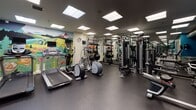 This fitness center offers a comprehensive workout environment designed to accommodate a wide range of exercise routines. The space is organized into distinct functional zones, making it easy to navigate between cardio, strength training, and free weight areas.

The cardio section features multiple treadmills and elliptical machines, each equipped with individual monitors for entertainment or workout tracking. A striking, colorful mural depicting a Pacific Northwest landscape — complete with a snow-capped mountain, rolling hills, and floral details — serves as a vibrant focal point along one wall, adding an energetic and inspiring ambiance to the space.

The strength training area is anchored by a substantial multi-function cable machine, accompanied by a weight bench and various resistance attachments, offering versatility for a full range of exercises. Full-length mirrors line the walls, visually expanding the space and allowing users to monitor their form throughout their workouts.

The free weight section includes a well-stocked dumbbell rack, kettlebells in multiple sizes, stability balls, and foam rollers neatly organized on shelving. A teal accent wall adds a bold visual contrast against the darker flooring and ceiling, lending the space a modern, professional aesthetic.

Recessed panel lighting throughout ensures consistent, bright illumination across all areas. The durable rubber flooring provides both safety and comfort underfoot, and the overall layout maximizes usability within the space.