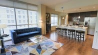 This contemporary single-floor residence showcases a seamlessly integrated open-concept layout that connects the living and kitchen areas into one cohesive, inviting space. Rich hardwood flooring flows throughout, anchoring the design with warmth and continuity.

The living area is generously proportioned, centered around a deep tufted navy-blue sofa and a geometric-patterned area rug in earth tones and blues. A sleek glass coffee table adds a modern touch, while a standing floor lamp provides ambient lighting. Large windows fitted with roller shades allow abundant natural light to fill the room while framing expansive urban views of the surrounding cityscape.

The kitchen is efficiently designed with dark cabinetry, stainless steel appliances, and a spacious light-colored island that doubles as a breakfast bar, accommodating several bar stools for casual dining or entertaining. Pendant lighting above the island adds a refined detail, complementing the modern aesthetic. A set of windows in the kitchen area mirrors those in the living space, reinforcing the bright and airy atmosphere throughout.

The overall ambiance is sleek and urban, blending contemporary furnishings with functional design to create a space that is both stylish and practical — ideal for modern city living.