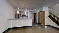 This two-story space showcases a modern, open-concept design with polished concrete floors that reflect natural light throughout. The kitchen serves as a central feature, equipped with stainless steel appliances including a refrigerator, microwave, and range. Warm wood cabinetry provides ample storage, while a granite-topped island offers additional workspace and defines the kitchen area. Pendant lighting hangs above both the island and kitchen workspace, complementing the recessed track lighting overhead.

The upper level is visible from the main floor, featuring a glass panel railing that maintains an open, airy feel between floors. Large windows fitted with horizontal blinds on the upper level allow generous natural light to filter down into the space below, enhancing the sense of openness and vertical scale. White-painted brick walls add a subtle textural contrast to the otherwise clean, neutral palette.

The layout includes multiple doorways leading to additional rooms, with dark wood door frames providing visual contrast against the light walls. The overall ambiance is contemporary and functional, combining industrial elements like concrete flooring with refined finishes such as granite countertops and pendant lighting.