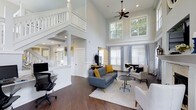 Located at 1100 Audubon Parc Drive in Cary, NC, this well-appointed clubhouse offers an inviting and functional environment for residents. The main gathering area immediately impresses with its dramatic double-height ceilings, which flood the space with natural light through large, generously sized windows framed by classic blue-and-white patterned curtains and white blinds. Views of mature trees just outside add a serene, nature-connected ambiance to the interior.

Rich, dark hardwood floors run throughout, providing a warm and polished foundation that complements the crisp white walls and trim. A ceiling fan anchors the upper expanse of the room, ensuring year-round comfort. The main lounge area is tastefully furnished with a modern gray sofa accented by mustard yellow throw pillows, complementary seating chairs, and a bistro-style table arrangement near the windows — ideal for casual conversation or a quiet retreat.

A dedicated workstation with a desk and ergonomic chair offers a practical space for residents who need to work or study. An upper-level interior balcony with white-painted railings overlooks the main floor, adding architectural character and visual depth. The overall design strikes a balance between classic craftsmanship and contemporary comfort, making this clubhouse a standout amenity for the Audubon Parc community.