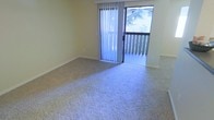 This single-floor space features a carpeted room finished with neutral-toned walls that create a clean, understated atmosphere. The room is equipped with plush wall-to-wall carpeting in a light beige tone, providing a comfortable and quiet underfoot feel throughout. A prominent sliding glass door serves as a key architectural feature, fitted with vertical blinds that allow for adjustable light control. The door opens directly onto a wood-railed balcony, where views of surrounding greenery, including mature trees, are visible. This connection to the outdoors enhances the sense of openness and brings natural light into the interior. The balcony's wooden railing adds a warm, natural contrast to the neutral interior palette. The room's layout is open and unobstructed, offering flexibility for various furniture arrangements. The overall ambiance is calm and functional, with the interplay of natural light filtering through the sliding door contributing to a bright and airy feel. The combination of soft carpeting, neutral walls, and outdoor access makes this a versatile and comfortable living space.