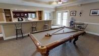 This single-floor recreational space is thoughtfully designed as a social and leisure hub, offering a welcoming atmosphere ideal for gathering and entertainment. The centerpiece of the room is a full-size billiards table, positioned on a patterned carpet that adds texture and visual interest to the space. Overhead, a ceiling fan with a light fixture provides both comfort and illumination, while a tray ceiling with crown molding lends an elegant architectural touch.

A built-in bar area lines one side of the room, featuring an open pass-through window, built-in shelving, and bar stool seating — ideal for casual socializing. The bar area is equipped for serving refreshments, making it a natural gathering point. On the opposite side of the room, a set of glass-paneled French doors floods the space with natural light and offers access to an outdoor area, enhancing the sense of openness.

Additional amenities include what appears to be a water or beverage station near the entrance, along with framed artwork on the walls that contribute to the room's polished, community-oriented character. The neutral color palette, combined with classic finishes and thoughtful furnishings, creates a comfortable and inviting environment suited for recreation and social events alike.